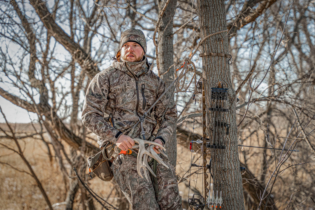 Man fully covered in ArcticShield Camo in a tree with a brown glass field behind him. Holding 2 antler sheds next to his hunting bow.