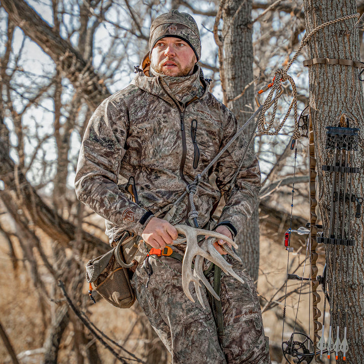 Hunter in the Prodigy Sentinel Pant holding deer antlers next to a tree stand in a forest setting.